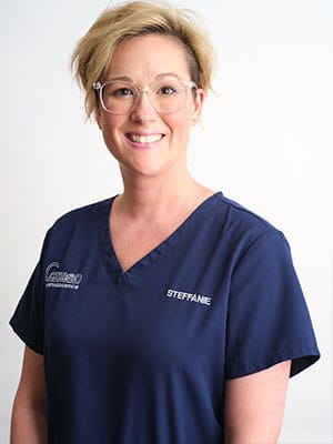 A blonde short haired woman in navy blue scrubs against a white background