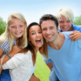 A mom and dad with brown hair and two kids with blonde hair smiling outside.