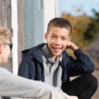 A boy with braces kneeling next to his friend outside