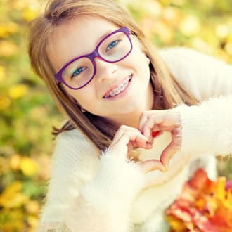 A girl with braces and purple glasses making a heart shape with ehr hand.