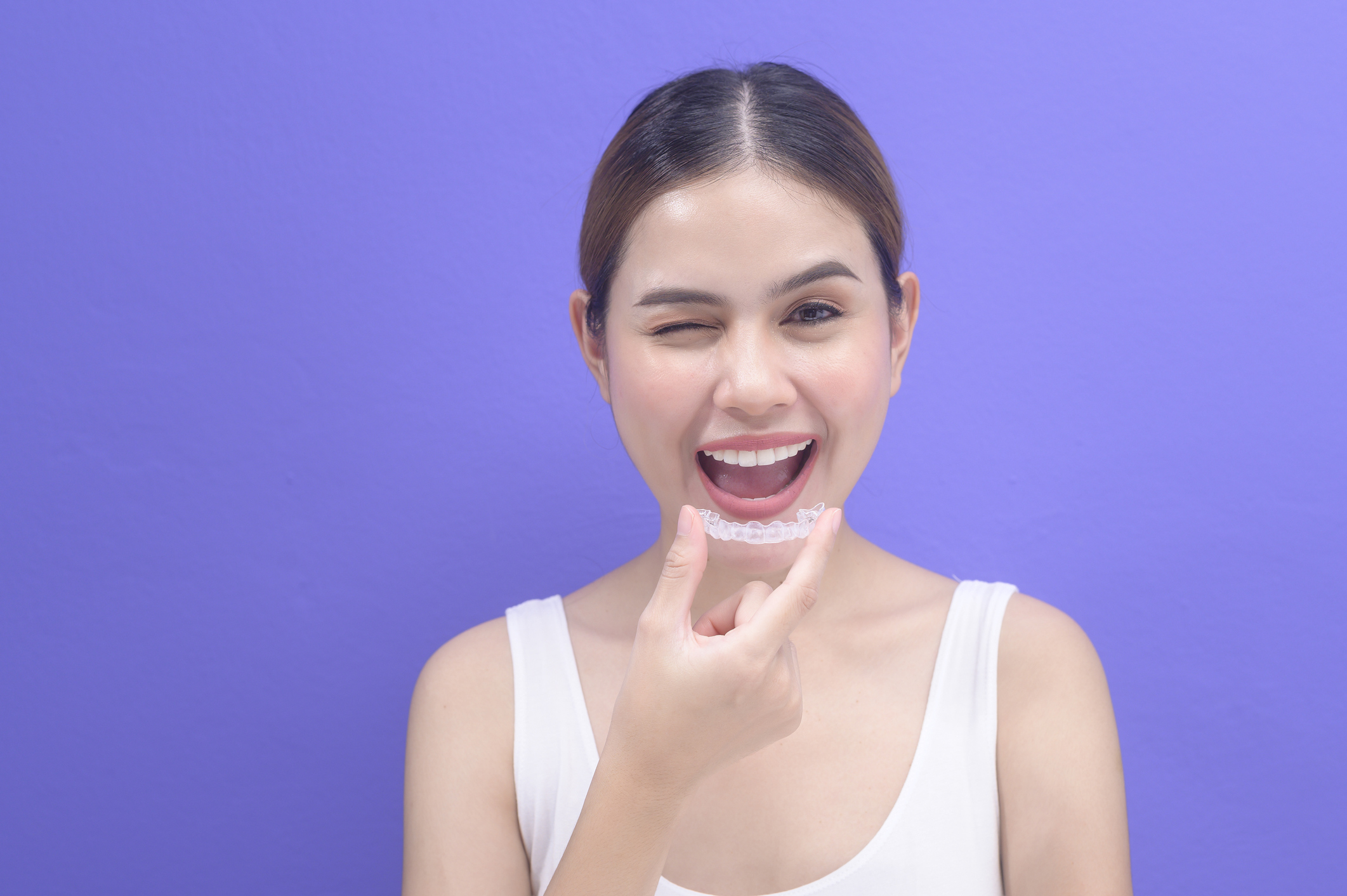 Winking dancer with short brown ponytail holding an invisalign aligner in a white tank top