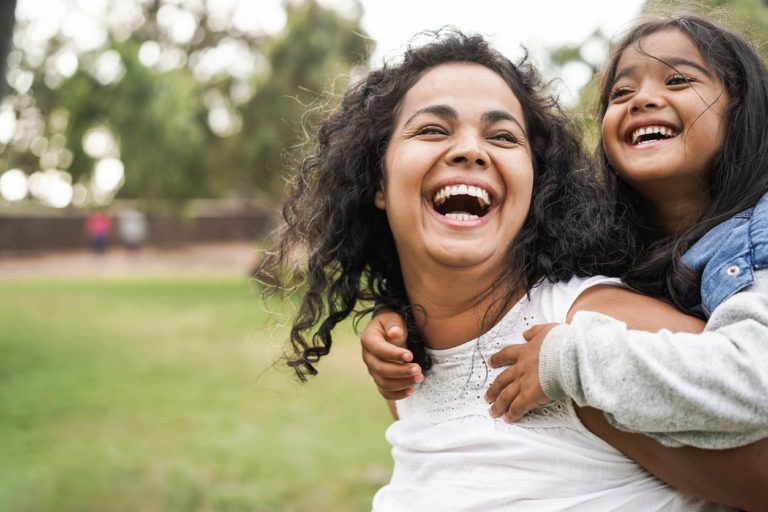 A mother laughing with her daughter as she carries her on her back through a park