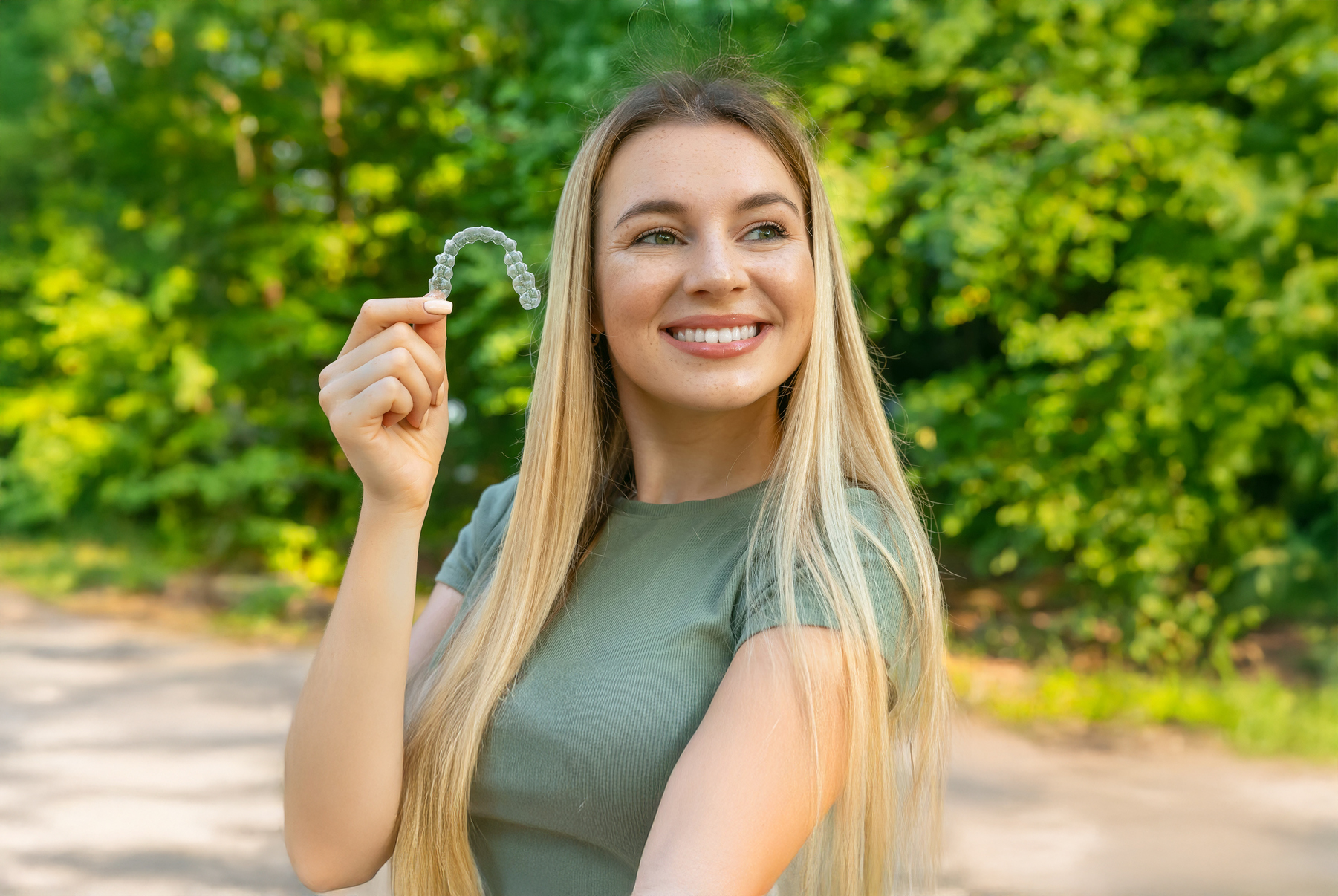 Woman with long blonde hair and an olive green shirt smiling and holding her invisalign aligner